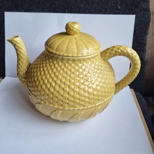 Peut inclure: Une théière en céramique jaune avec un motif de panier tressé. La théière a un bec courbé, une anse arrondie et un couvercle avec un petit bouton. La théière est de couleur jaune clair.