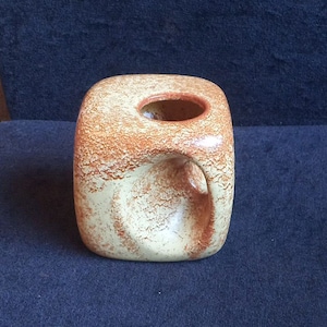 Puede incluir: Un jarrón cuadrado de cerámica con superficie texturizada. El jarrón tiene una base beige claro con detalles en naranja-marrón y una abertura circular en la parte superior. Una abertura curva está en un lado.