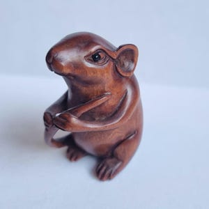 May include: A small, hand-carved wooden figurine of a mouse with a smooth, polished dark brown finish. The mouse is depicted in a seated position, holding a thin, stick-like object with its front paws, featuring detailed ears, eyes, and feet.