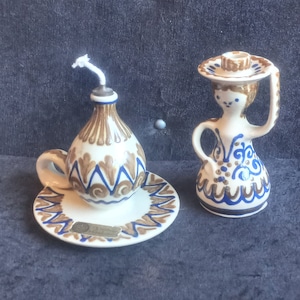 May include: A ceramic oil lamp and a decorative figurine. The oil lamp is white with brown and blue patterns, a handle, and a wick. The figurine is a woman holding a hat, with similar blue and brown patterns.