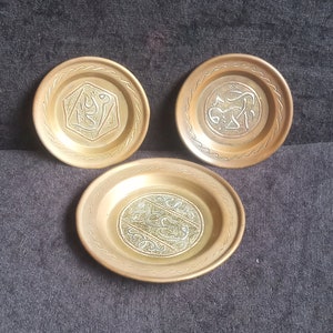 May include: Three antique brass plates with intricate engraved Arabic calligraphy. Each plate has a circular design with a detailed border. The plates are arranged on a dark gray surface.