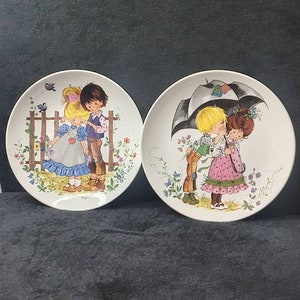 Può includere: Due piatti decorativi con scene romantiche. Un piatto raffigura una coppia vicino a una staccionata di legno, l'altro una coppia sotto un ombrello. Entrambi i piatti presentano decorazioni floreali e a farfalla, con un'estetica vintage.