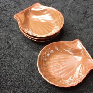 May include: Four copper-orange seashell-shaped dishes are shown, with a glossy finish. The dishes are stacked, with one separate. The background is a dark, textured fabric. Each dish is designed to resemble a seashell.
