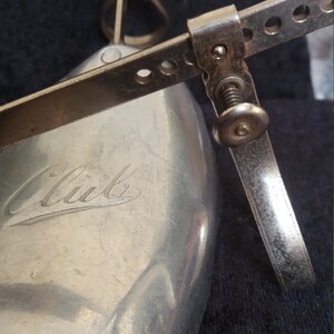 May include: Close-up of a vintage shoe stretcher made of silver-toned metal. The stretcher has a curved shoe form with the word "Club" engraved on it. Adjustable metal arms and a screw mechanism are visible.