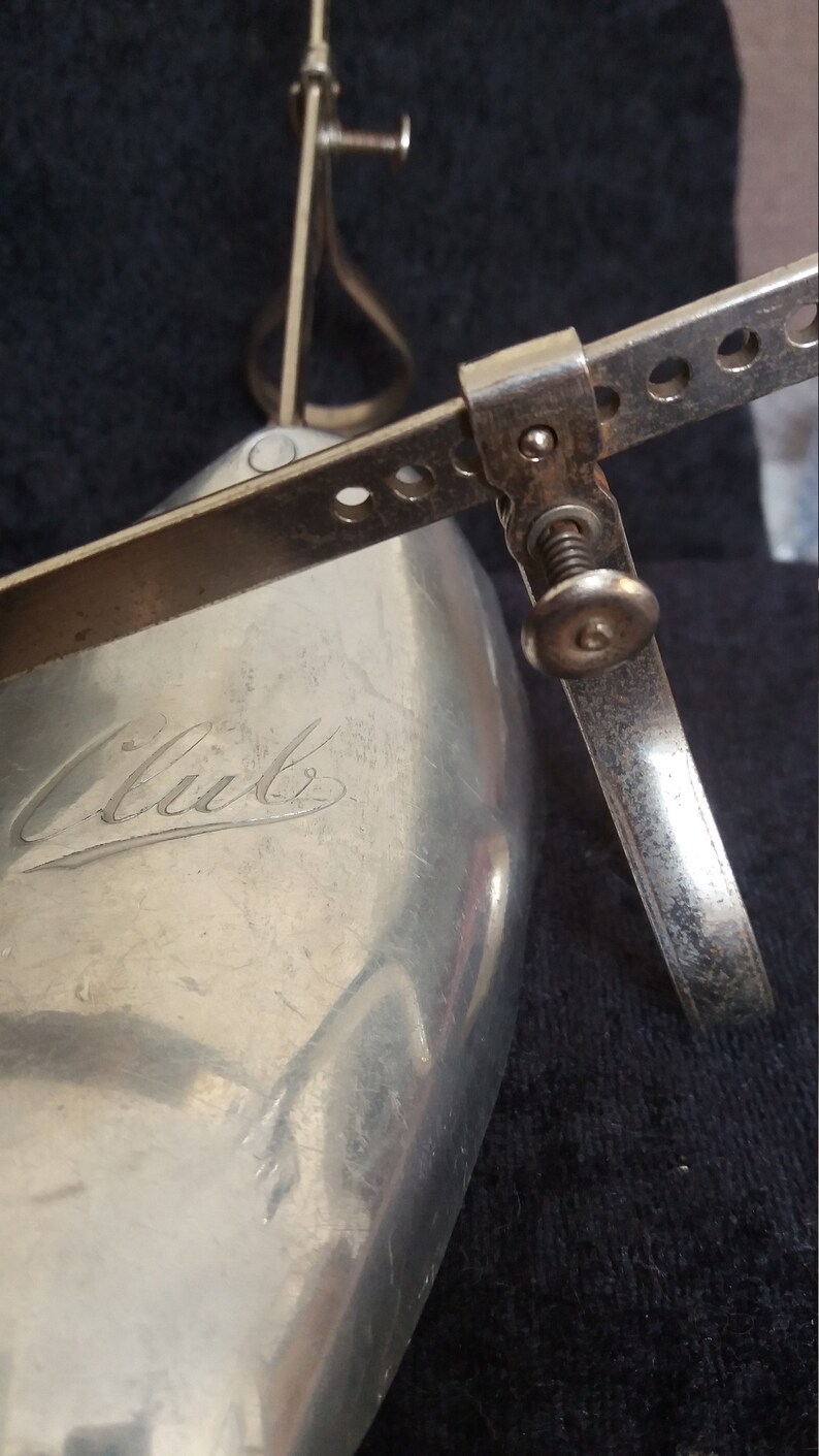 May include: Close-up of a vintage shoe stretcher made of silver-toned metal. The stretcher has a curved shoe form with the word "Club" engraved on it. Adjustable metal arms and a screw mechanism are visible.