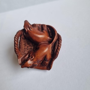 May include: A small, circular, brown wooden carving featuring two rats positioned within a rope-like border. The rats are sculpted with smooth surfaces and dark, inlaid eyes, resting on a textured base that resembles woven fabric or straw.