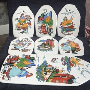 Może przedstawiać: Zestaw siedmiu białych ceramicznych półmisków i desek do krojenia z kolorowymi ilustracjami sztuki ludowej. Projekty przedstawiają sceny wędkarskie, gotowania i życia nadmorskiego. Zestaw zawiera długą prostokątną deskę i sześć mniejszych, unikalnych elementów.