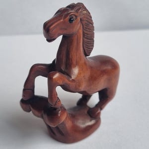 May include: A small, hand-carved wooden figurine of a horse in a rearing position. The sculpture features a smooth, polished reddish-brown finish, detailed mane texture, and a solid base that supports the horse as it stands on its hind legs.