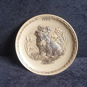 Puede incluir: Plato decorativo beige con un relieve de un perro sosteniendo una flor en la boca, rodeado de flores y hierba. El año "1981" está encima del perro. El plato tiene un borde decorativo.