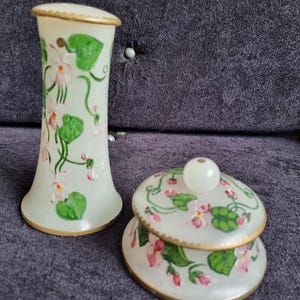 Puede incluir: Un conjunto decorativo que presenta un jarrón y un tarro con tapa. Ambas piezas son blancas con ribetes dorados y diseños florales pintados a mano en rosa y verde. El tarro tiene un pomo blanco en la parte superior.