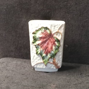 Peut inclure: Un vase en céramique blanc avec une surface texturée et un motif de feuille peint. La feuille est principalement rouge et verte avec des accents dorés. Le vase est rectangulaire et a une base carrée.