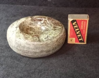 Puede incluir: Un cenicero redondo de piedra con una depresión central y un exterior texturizado y en capas. El cenicero es de color verde y marrón moteado. Una caja de cerillas "VELVET" está al lado del cenicero.