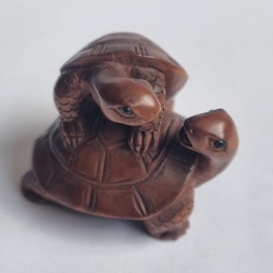 Op de afbeelding: Een donkerbruin uitgesneden houten beeldje van twee schildpadden, waarbij de ene schildpad boven op de andere zit. Beide schildpadden hebben gedetailleerde schildpatronen, een getextureerde huid op de ledematen en kleine donkere ogen, gefotografeerd van bovenaf.