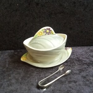 May include: A vintage, pale blue ceramic sugar bowl with a lid, on a matching saucer. The lid and saucer feature yellow and purple floral details. Silver tongs are positioned in front of the bowl.