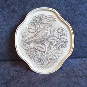 Peut inclure: Assiette décorative blanche cassé, à bord festonné, avec un relief en relief d'un oiseau perché sur une branche fleurie. L'oiseau chante, le bec ouvert. L'assiette a une fine bordure bleue.
