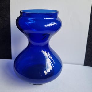 Puede incluir: Un jarrón de vidrio azul cobalto con una forma única de reloj de arena. El jarrón tiene una base ancha y una parte superior acampanada, con una sección media más estrecha. El interior del jarrón es de un tono azul más claro. Adecuado para la decoración del hogar.