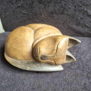 May include: A wooden cat sculpture in a curled-up position. The cat is crafted from light brown wood, showcasing the natural grain and texture. The ears are pointed, and the tail is wrapped around the body. The sculpture has a smooth finish and rests on a grey base.