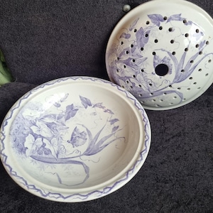 Könnte beinhalten: Zwei weiße Keramik-Servierschalen mit floralem Design in Lilatönen. Eine Schale ist flach, die andere ein Deckel mit Löchern. Das florale Muster umfasst Blätter und Schmetterlinge.