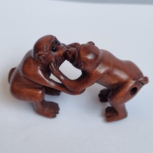 May include: A brown carved wooden netsuke sculpture depicting two monkeys facing each other with their hands touching. The monkeys have smooth, polished surfaces and detailed facial features, with a small circular hole visible on the side.