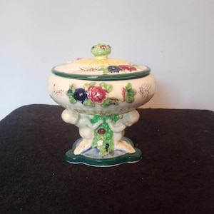 Könnte beinhalten: Eine Keramik-Schmuckschatulle mit Deckel. Die Schale ist cremefarben mit bunten Blumenmustern und grünen Verzierungen. Der Sockel wird von zwei cherubartigen Figuren und einem grünen Sockel getragen. Der Deckel hat einen kleinen grünen Knopf.