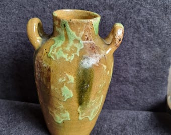 Pode incluir: Um pequeno vaso de cerâmica artesanal com duas alças. O vaso tem uma cor de base amarelo-castanho com detalhes verdes e castanhos. O vaso é provavelmente usado para fins decorativos.