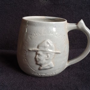 Peut inclure: Mug en céramique blanc cassé moucheté avec une grande anse. La tasse présente un motif en relief d'un homme portant un chapeau et les mots "BADEN-POWELL LICENSE 1857-1957".