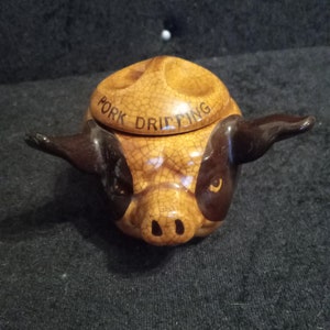May include: A ceramic pig-shaped container with a lid, in brown and tan. The lid is inscribed with the words "Pork Dripping". The container has dark brown ears and snout, and is designed to resemble a pig's face.
