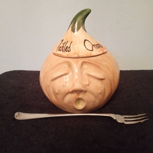 May include: A ceramic pickled onion jar with a crying face design, featuring a green stem and the words "Pickled Onions." A silver fork rests in front of the jar. The jar is tan and cream colored.
