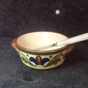 May include: A small, hand-painted ceramic bowl with a cream interior and a decorative floral pattern in green, blue, and red. The bowl has brown handles and a white-handled knife rests inside.