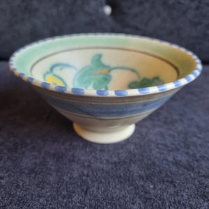 May include: A ceramic bowl with a footed base, featuring a hand-painted design. The rim is bordered with blue dots and a green band. The interior displays green and yellow floral motifs against a cream background. The bowl has a diameter of approximately 6 inches.