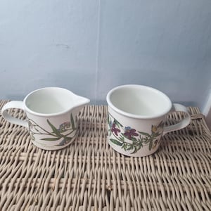 May include: Two white ceramic items with floral patterns. A small creamer with a handle and spout, and a mug with a handle. Both feature a botanical design of purple, blue, and yellow flowers with green leaves.