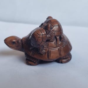 May include: A brown carved wooden figurine depicting a large turtle with a smaller turtle resting on its shell. The sculpture features detailed textures on the shells and limbs, with a smooth, polished finish throughout the entire wooden surface.