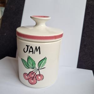 以下が含まれることがあります： クリーム色のセラミックジャムジャー。蓋と赤いストライプが付いています。「JAM」の黒い文字と、緑の葉が付いたサクランボの房が描かれています。ヴィンテージのキッチン用品です。