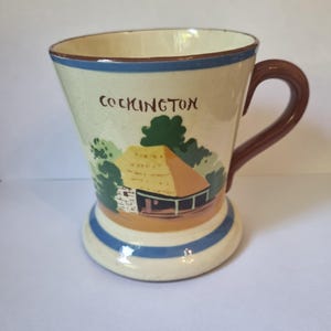 May include: A vintage ceramic mug with a brown handle and a flared shape. The mug features a painted scene of a cottage with a thatched roof, green trees, and the word "COCKINGTON" in brown lettering. Blue and brown bands decorate the base.