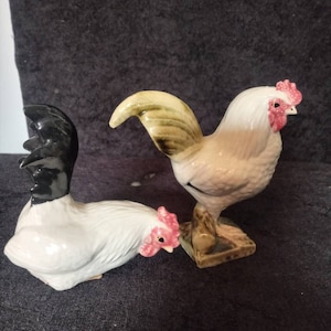 May include: Two ceramic rooster figurines. One is standing upright, the other is lying down. Both have white bodies, red combs, and black tail feathers. The standing rooster is on a square base.