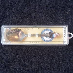 May include: A silver spoon in a clear plastic case. The spoon features a detailed handle with a Beatrix Potter design, including a small figure and a circular emblem. The spoon is displayed on a yellow background.
