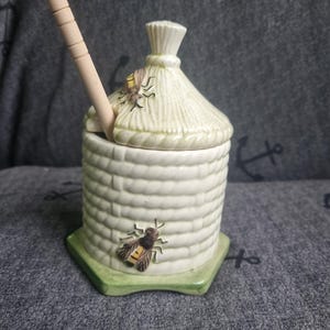 May include: A ceramic honey pot with a beehive design. The pot is off-white with a textured surface and a straw-like lid. A wooden honey dipper rests inside. Two decorative bees are attached to the pot. The pot sits on a green hexagonal base.