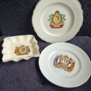 May include: Three vintage white ceramic plates with gold trim. One plate features a royal emblem with a portrait, another has a scalloped edge and a similar emblem, and the third displays portraits and flags.