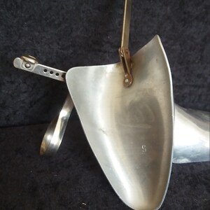 May include: A vintage shoe stretcher made of silver-colored metal. The device has a curved, shovel-like form with an adjustable mechanism and a handle. The stretcher is designed to expand the size of shoes. The background is dark.