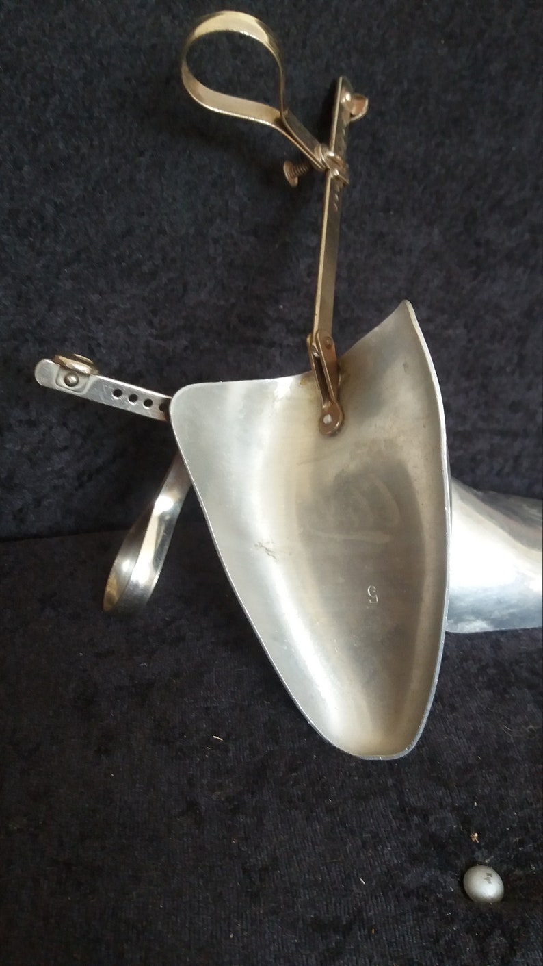 May include: A vintage shoe stretcher made of silver-colored metal. The device has a curved, shovel-like form with an adjustable mechanism and a handle. The stretcher is designed to expand the size of shoes. The background is dark.