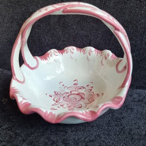 May include: A white ceramic basket with a handle, featuring a scalloped rim and a floral design in shades of pink. The handle and rim are accented with pink detailing. The basket is likely for decorative use.