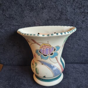 May include: Cream-colored ceramic vase with a flared rim and a round base. The vase is decorated with a floral design in shades of blue, purple, green, and pink. The rim has small blue dots. A decorative piece for the home.