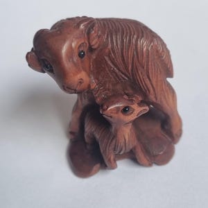 May include: A brown carved wooden figurine depicting a large ram standing over a smaller lamb. The sculpture features detailed textures representing the animal fur, with dark inlaid eyes on both figures, capturing a protective stance.