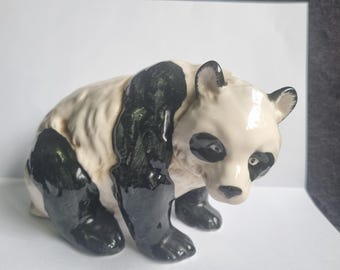 Op de afbeelding: Een keramieken panda beeldje met zwart-witte kleuren. De panda zit, met zwarte poten en armen, en zwarte vlekken rond de ogen en oren. Het beeldje heeft een glanzende afwerking.