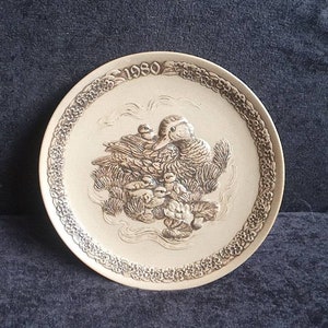 Puede incluir: Un plato decorativo redondo, de color crema, con un diseño en relieve. El plato presenta una escena familiar de patos con el año "1980" en la parte superior. El borde tiene un patrón floral. El plato está sobre una superficie azul oscuro.