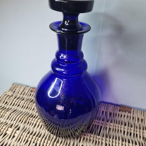 Puede incluir: Una jarra de vidrio azul cobalto con un tapón negro. La jarra tiene una base redondeada y un cuello largo. El tapón es plano y redondo. La jarra está sobre una superficie de mimbre tejido.