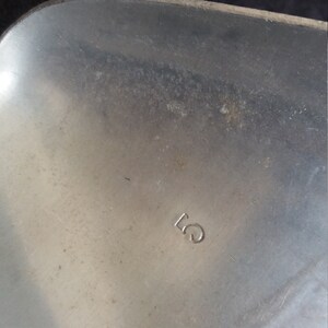 May include: Close-up of a silver metal object with the number "5" stamped on the surface. The metal has a brushed texture and some minor imperfections. The object appears to be a part of a larger item.