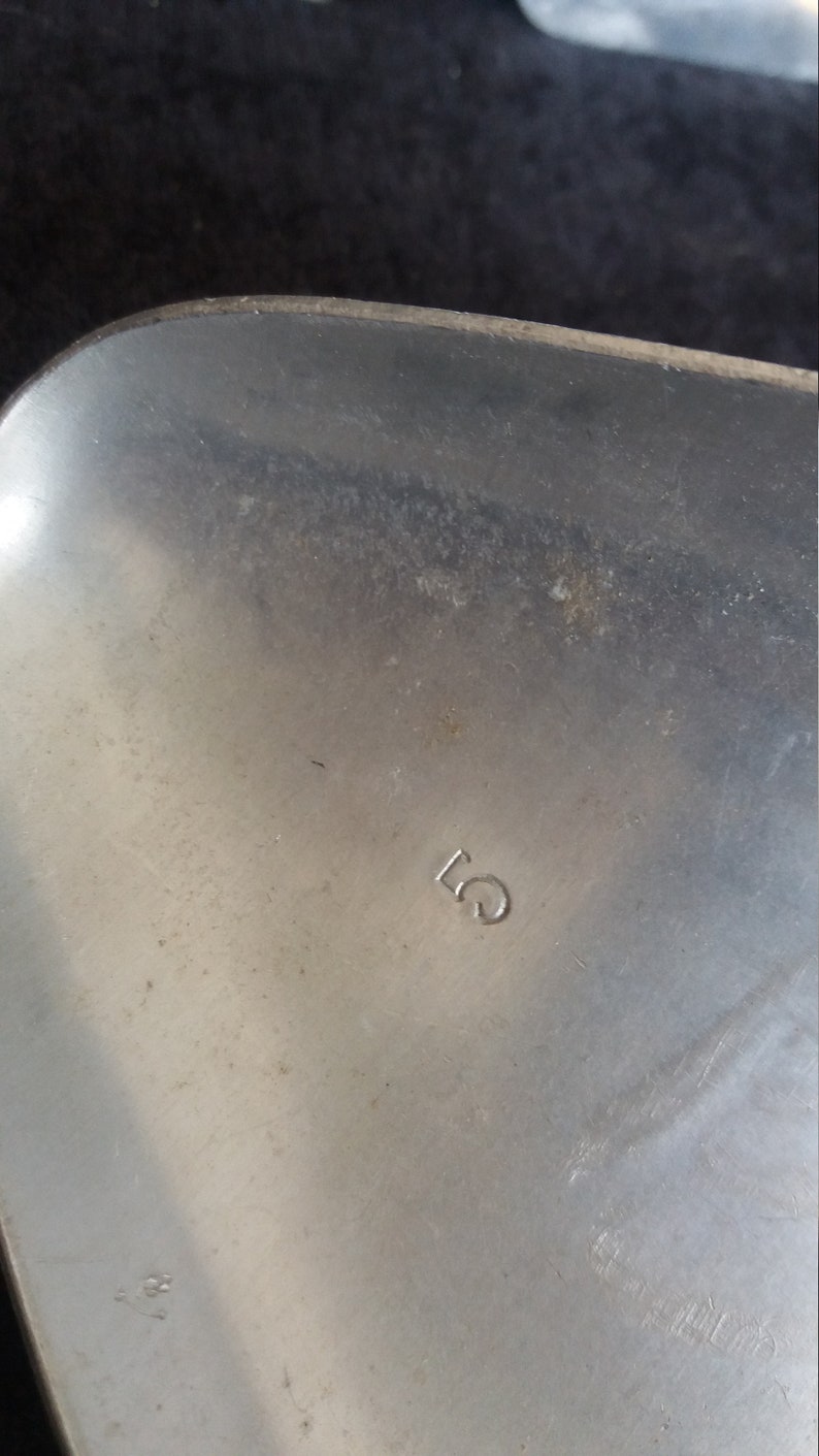 May include: Close-up of a silver metal object with the number "5" stamped on the surface. The metal has a brushed texture and some minor imperfections. The object appears to be a part of a larger item.
