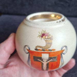 May include: A small, round, off-white ceramic container with a gold rim. The container features a detailed illustration of a cornucopia, a cross, and a figure. The design includes red, orange, and blue accents. The container is held in a hand.