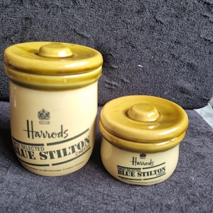 May include: Two ceramic Harrods Blue Stilton cheese jars. The jars are cream-colored with a yellow-brown lid and a matching rim. The Harrods logo and text are printed on the front of the jars. The jars are cylindrical and have a small knob on the lid.